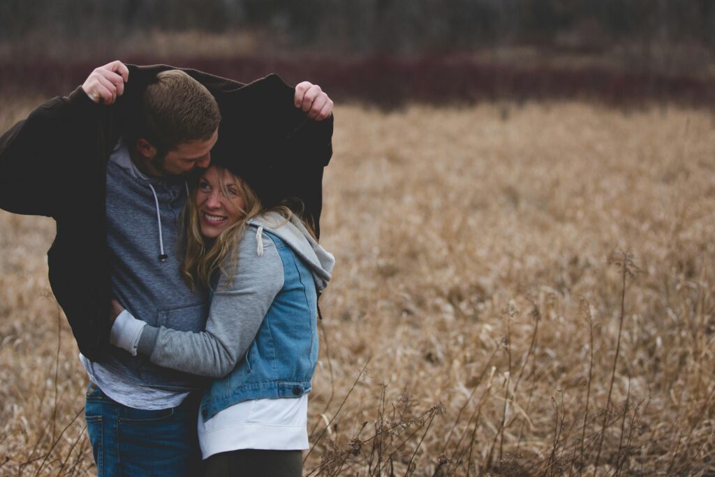 A couple shares a warm embrace under a coat in a rustic autumn field, expressing love and togetherness.